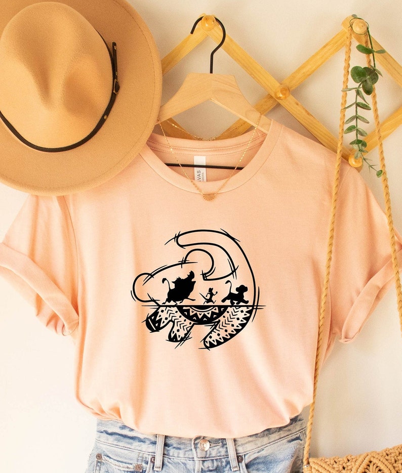 May include: A peach-colored t-shirt with a black silhouette of the Lion King's "circle of life" symbol. The symbol features Simba, Nala, and Timon running across the savanna.