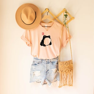 May include: A light pink t-shirt with a black frog illustration and a red stamp. The shirt is hanging on a wooden hanger with a brown hat and a straw bag.