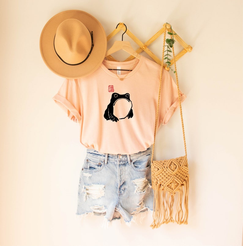 May include: A light pink t-shirt with a black frog illustration and a red stamp. The shirt is hanging on a wooden hanger with a brown hat and a straw bag.