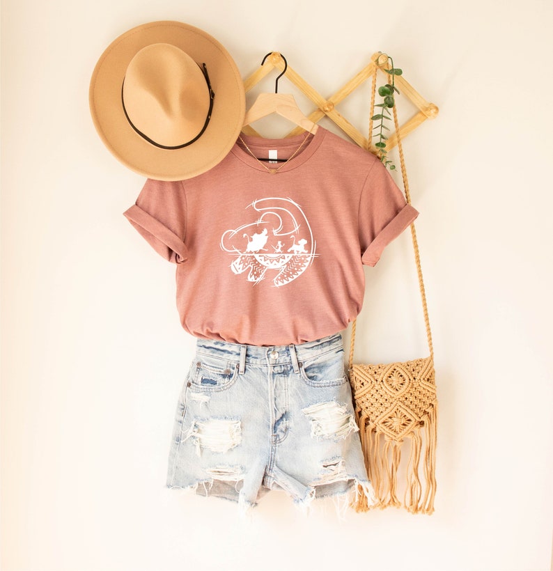 May include: A dusty rose t-shirt with a white graphic of the Lion King silhouette. The shirt is hanging on a wooden hanger with a brown hat and a straw bag.