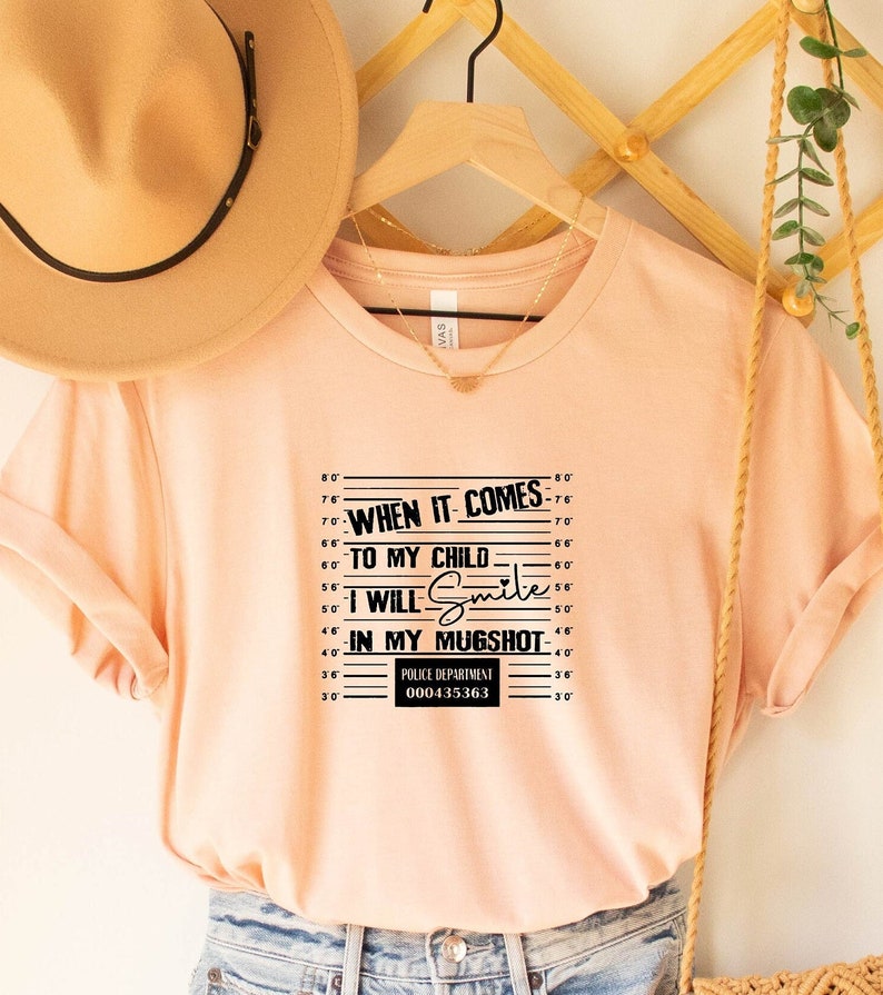 Może przedstawiać: R&oacute;żowy t-shirt z czarnym nadrukiem przedstawiającym zdjęcie policyjne. Nadruk zawiera tabelę wzrostu, tekst "Kiedy chodzi o moje dziecko, będę się uśmiechać na zdjęciu policyjnym" oraz numer departamentu policji.