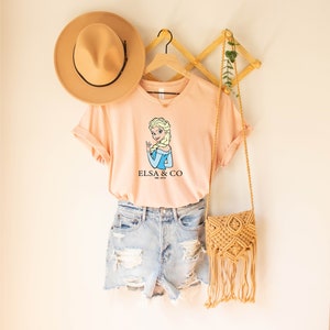 May include: A peach-colored t-shirt with a cartoon image of Elsa from Frozen. The shirt has the text "Elsa & Co. Est. 2013" printed on it. The shirt is hanging on a wooden hanger with a straw hat and a woven bag.