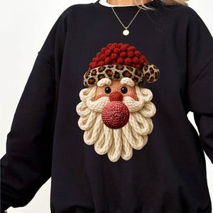 Peut inclure: Sweat-shirt noir avec un motif de Père Noël. Le visage du Père Noël est orné d'un bonnet rouge, d'une bordure léopard, d'un visage tricoté et d'un grand nez rouge. La barbe est faite de corde crème.