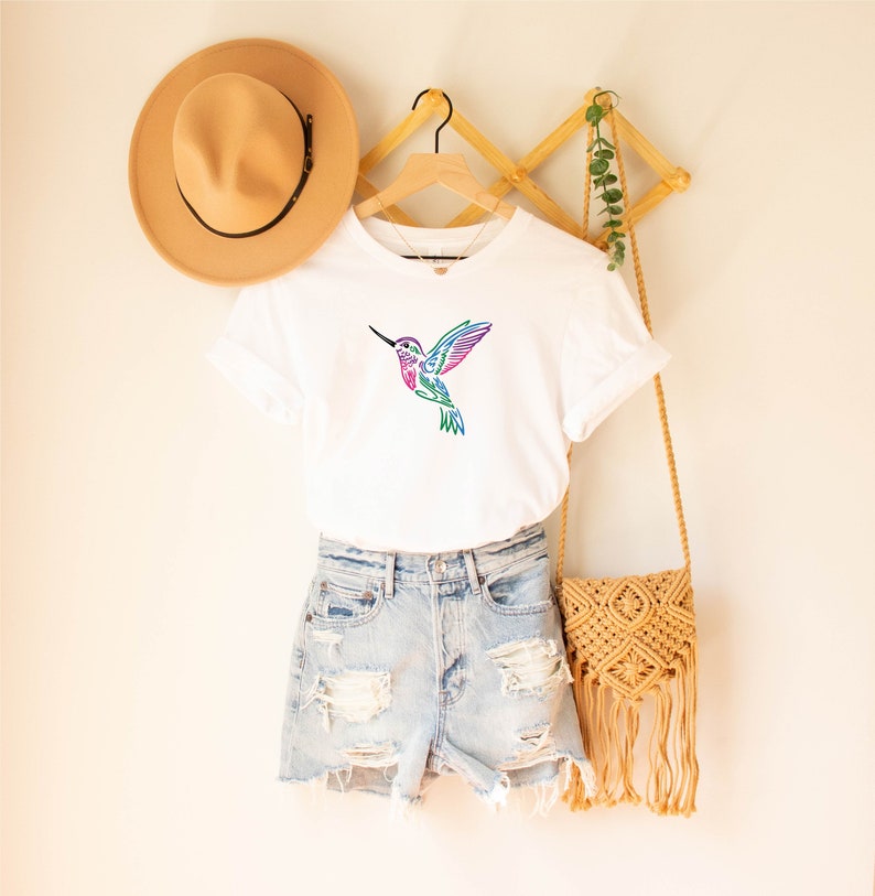May include: White t-shirt with a colorful hummingbird graphic. The hummingbird is stylized and has a rainbow color scheme. The shirt is displayed on a hanger with a straw hat and a woven bag.