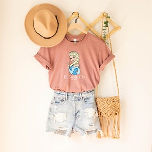 May include: A pink t-shirt with a cartoon image of Elsa from Frozen and the text "Elsa & Co.". The shirt is hanging on a wooden hanger and is paired with denim shorts and a straw bag.
