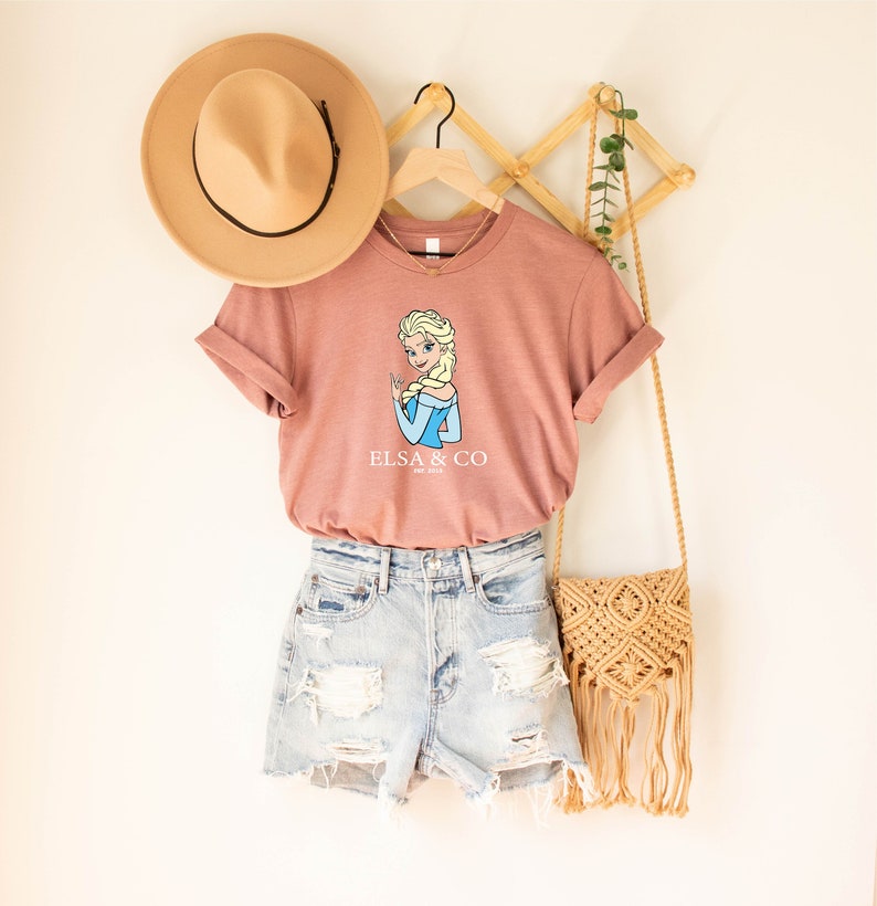 May include: A pink t-shirt with a cartoon image of Elsa from Frozen and the text "Elsa & Co.". The shirt is hanging on a wooden hanger and is paired with denim shorts and a straw bag.