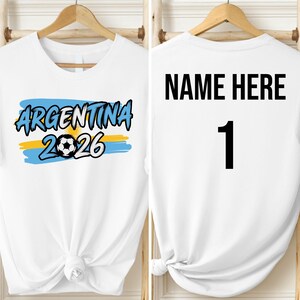 May include: White t-shirt with the word "ARGENTINA" in blue, a soccer ball, and the number "2026" in black. The back of the shirt has "NAME HERE" and the number "1" in black. The shirt is hanging on a wooden hanger.