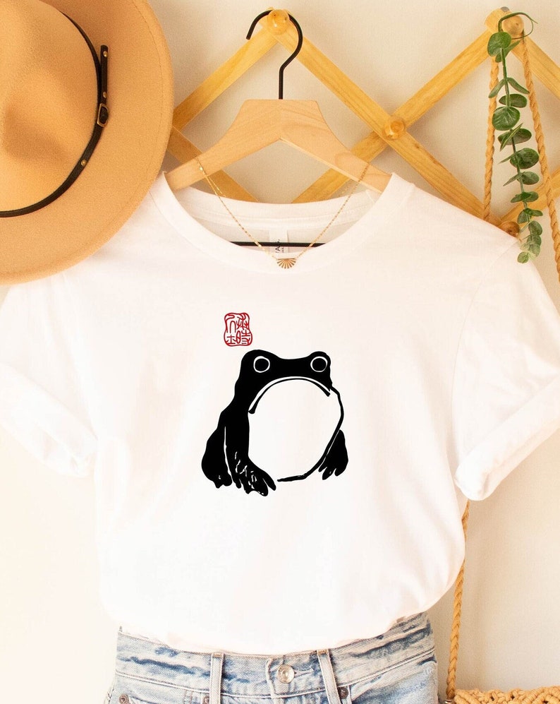May include: White t-shirt with a black silhouette of a frog and a red stamp above it.