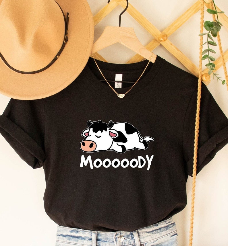 May include: Black t-shirt featuring a cartoon cow with black and white spots, a pink nose, and closed eyes. The word "MOOOOODY" is printed in white below the cow. A tan hat and gold necklace are visible in the background.