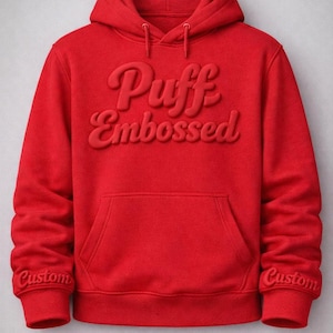 Op de afbeelding: Een felrode hoodie met een voorzak en een capuchon. De woorden "Puff Embossed" staan in een verhoogd, puffy lettertype. De manchetten van de mouwen hebben het woord "Custom" geborduurd.