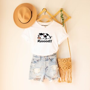 May include: White t-shirt with a cartoon cow graphic and the word "MoooooDy" in black. The shirt is paired with denim shorts, a tan hat, and a woven crossbody bag. The items are displayed on a wooden hanger.