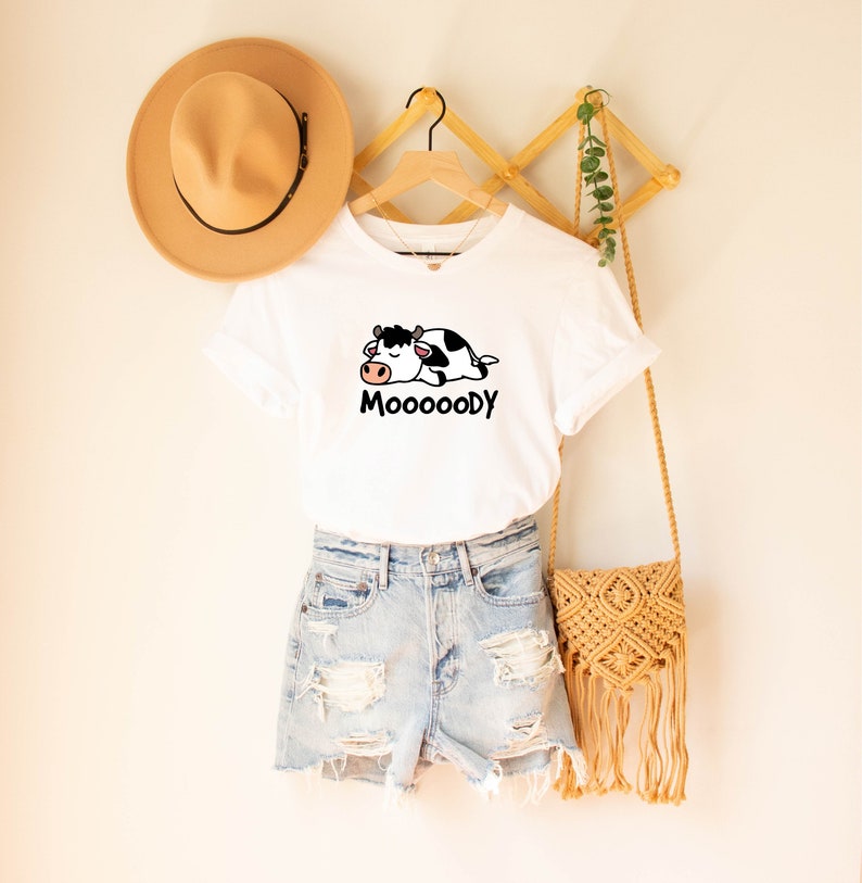 May include: White t-shirt with a cartoon cow graphic and the word "MoooooDy" in black. The shirt is paired with denim shorts, a tan hat, and a woven crossbody bag. The items are displayed on a wooden hanger.