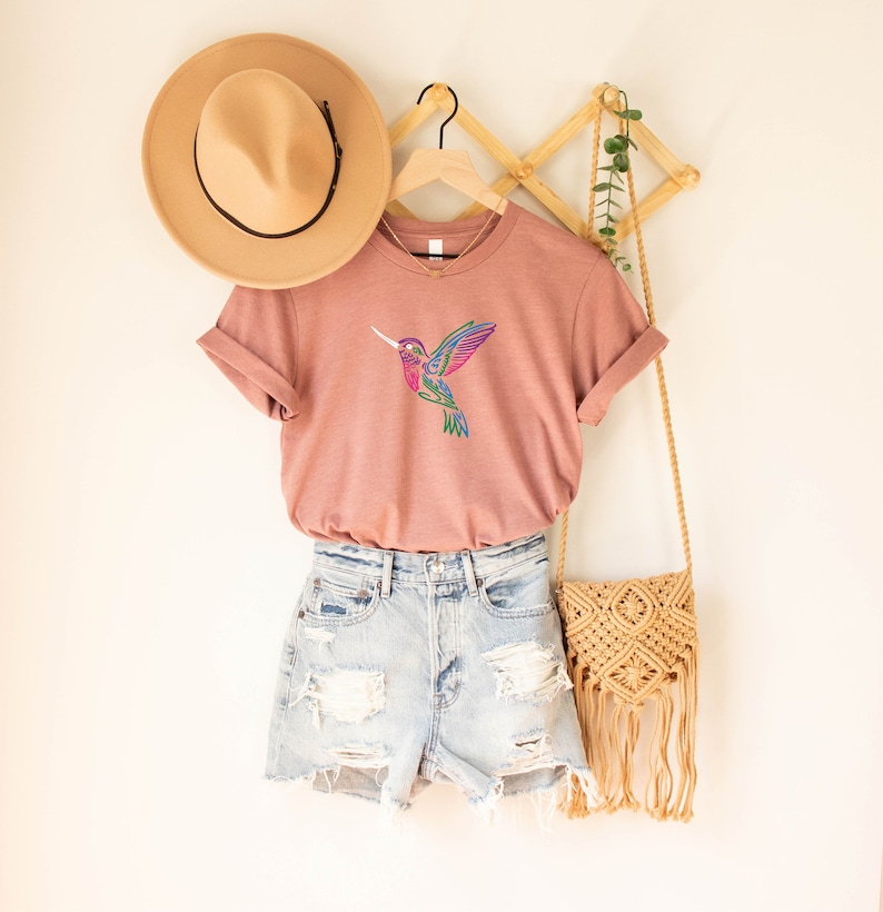 May include: A dusty rose t-shirt with a colorful hummingbird graphic. The shirt is hanging on a wooden hanger with a straw hat and a woven bag.