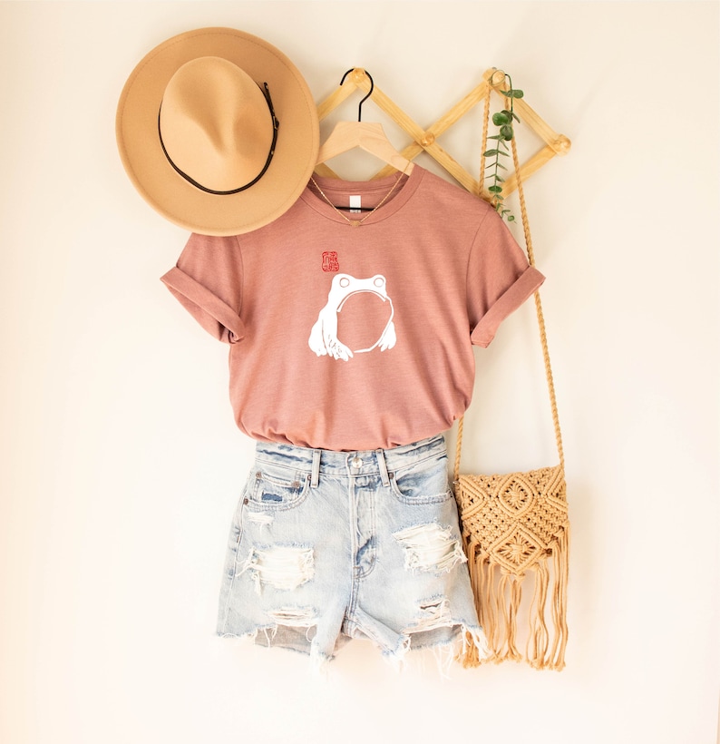 May include: A mauve t-shirt with a white frog graphic and a red stamp. The shirt is hanging on a wooden rack with a straw hat and a woven bag.
