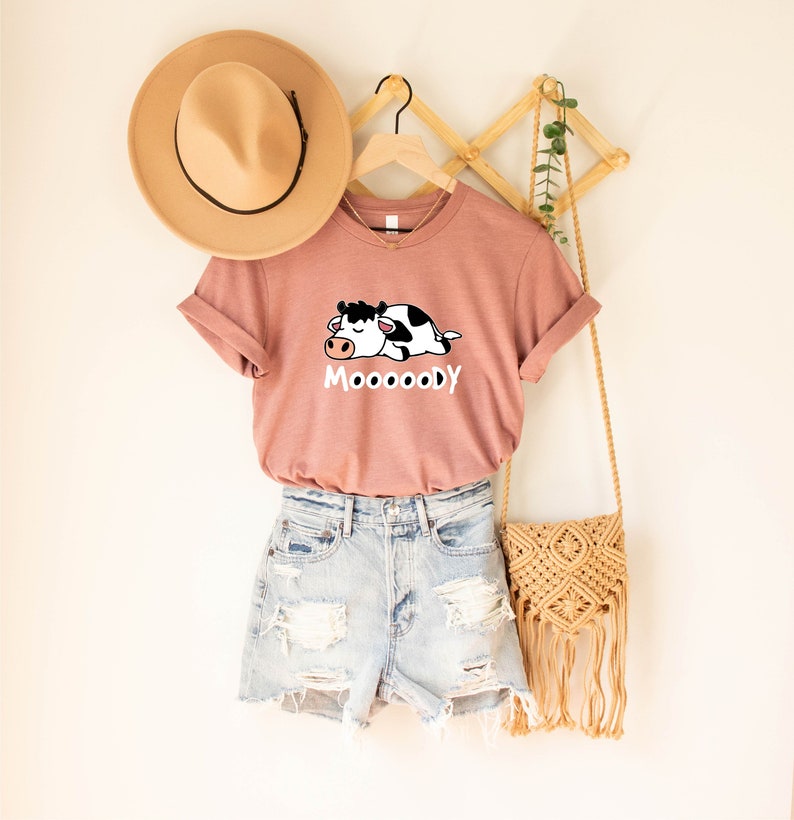 May include: A dusty rose t-shirt with a graphic of a sleeping black and white cow and the word "MoooooDY" in white. The shirt is paired with light blue denim shorts and a tan hat. A macrame bag hangs nearby.