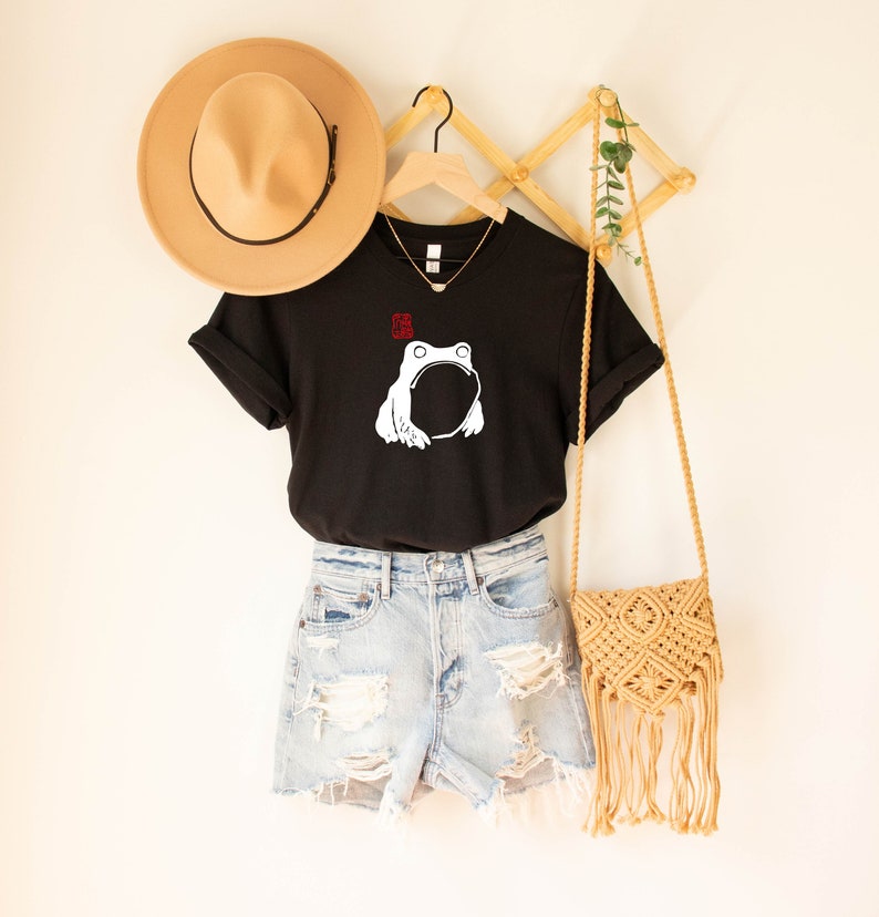 May include: Black t-shirt with a white graphic of a frog. The shirt is hanging on a wooden hanger with a straw hat and a woven bag.
