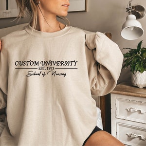 Pode incluir: Uma camisa de moletom bege com o texto "CUSTOM UNIVERSITY - EST. 1973 - School of Nursing" impresso na frente.