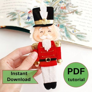 Puede incluir: Patrón de adorno navideño de fieltro de un Cascanueces soldado con uniforme rojo y dorado, barba blanca y sombrero negro. El patrón incluye un tutorial PDF para hacer el adorno.