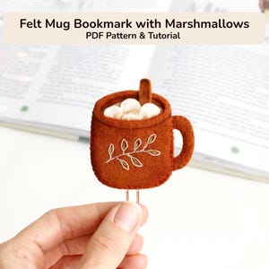May include: A brown felt mug bookmark featuring white marshmallow shapes inside and a stitched leaf design on the front. The text Felt Mug Bookmark with Marshmallows PDF Pattern & Tutorial is displayed above the mug shape.