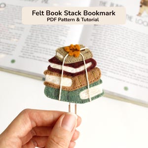 May include: A felt book stack bookmark featuring four stacked books in shades of tan, maroon, brown, and green, tied together with white string and topped with a small orange flower, attached to a metal paperclip. Text reads Felt Book Stack Bookmark.