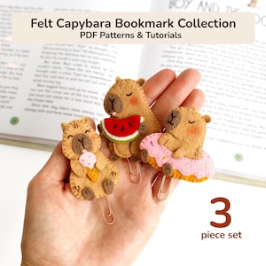 Könnte beinhalten: Ein Set aus drei Filz-Lesezeichen in Form von Capybaras an kupferfarbenen Büroklammern. Ein Capybara hält ein rosa Eis, eines eine Wassermelone und eines sitzt in einem rosa glasierten Donut. Der Text lautet Felt Capybara Bookmark Collection.