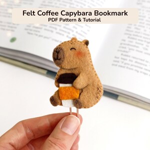Peut inclure: Un marque-page en feutrine beige représentant un capybara avec un nez brodé marron, des joues roses et une tasse de café avec un couvercle marron, un milieu orange et une base blanche. Le texte "Felt Coffee Capybara Bookmark PDF Pattern & Tutorial" est visible.