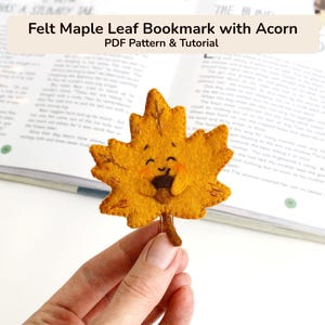 May include: A yellow felt maple leaf bookmark featuring a small brown acorn held in the centre. The leaf has embroidered details, a stitched border, and a smiling face with rosy cheeks. The text Felt Maple Leaf Bookmark with Acorn PDF Pattern & Tutorial is visible at the top.