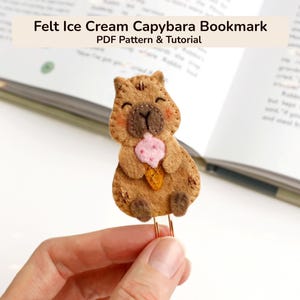 May include: A felt capybara bookmark with an ice cream cone. The capybara is brown with a pink and orange ice cream cone. The bookmark is attached to a copper-coloured paper clip. Text at the top reads "Felt Ice Cream Capybara Bookmark PDF Pattern & Tutorial."