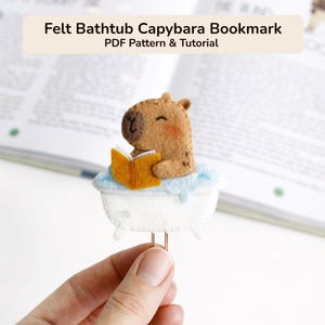May include: A felt bookmark featuring a brown capybara sitting in a white bathtub filled with light blue bubbles, holding an open yellow book. The text Felt Bathtub Capybara Bookmark PDF Pattern & Tutorial is visible at the top of the image.
