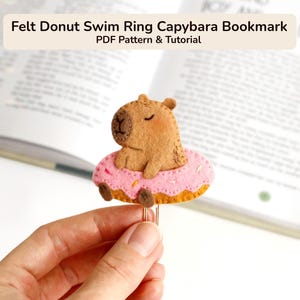 Puede incluir: Un marcapáginas de fieltro con un capibara marrón sentado dentro de un flotador con forma de dónut rosa con virutas de colores. El capibara tiene detalles faciales cosidos y los ojos cerrados. Unido a un clip metálico para papel.