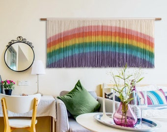 Puede incluir: Un tapiz de pared de macramé con los colores del arcoíris. La obra de arte presenta un degradado de colores, incluyendo rojo, naranja, amarillo, verde, azul y morado, dispuestos en forma de arco. El tapiz está suspendido de un listón de madera.
