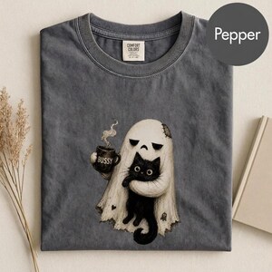 May include: A pepper-colored t-shirt featuring a graphic of a white ghost holding a black cat and a mug with the text BUSSY. The shirt has a crew neckline and a Comfort Colors label at the inner neck. The graphic includes steam rising from the mug.