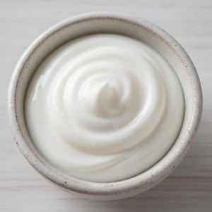 May include: A close-up shot of a small, round, ceramic bowl filled with white, creamy yogurt. The yogurt has a smooth, swirled texture. The bowl is light gray with a speckled rim, set against a light wooden surface.