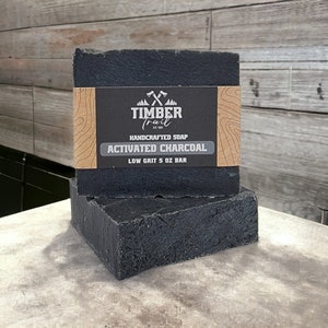 Może przedstawiać: Dwa czarne mydła z węglem aktywnym z brązową etykietą z napisem "Timber Trail Est. 1993 Ręcznie robione mydło Węgiel aktywny Niskie ziarno 5 oz Słup".