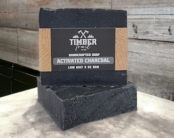 Op de afbeelding: Twee zwarte zeepstaven met geactiveerde houtskool en een bruin etiket met de tekst "Timber Trail Est. 1993 Handgemaakte zeep Geactiveerde houtskool Fijne korrel 5 oz Staaf".