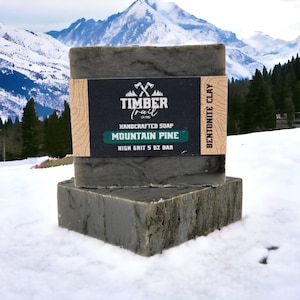 May include: A bar of dark gray soap with a green and black label that reads "Timber Trail Est. 2015 Handcrafted Soap Mountain Pine High Grit 5 oz Bar Bentonite Clay". The soap is sitting on a snowy surface with a mountain range in the background.