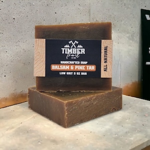 May include: Two bars of brown soap stacked on top of each other. The top bar has a label that reads "TIMBER Trail HANDCRAFTED SOAP BALSAM & PINE TAR LOW GRIT 5 OZ BAR ALL NATURAL".