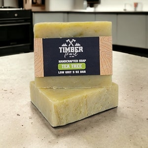 Op de afbeelding: Twee handgemaakte theeboomzeepblokken. De zeep is lichtgroen met een textuuroppervlak. Een zwart etiket met het Timber Trail-logo en "Tea Tree" is zichtbaar. Elk blok weegt 142 gram.