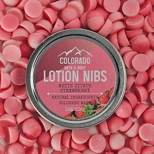 May include: A silver tin of white citrus and strawberry lotion nibs from Colorado Bath & Body. The tin is surrounded by pink lotion nibs.