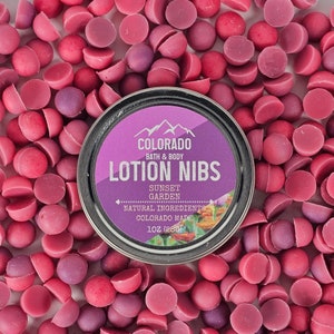 May include: A silver tin with a purple label that reads "COLORADO BATH & BODY LOTION NIBS SUNSET GARDEN NATURAL INGREDIENTS COLORADO MADE 1 OZ (280)". The tin is surrounded by small, pink and purple, half-sphere shaped bath products.