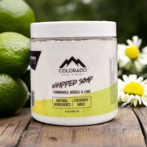 May include: A white jar of whipped soap with a green label. The label says "Colorado Bath & Body" and "Whipped Soap" in black text. The label also says "Chamomile, Neroli & Lime" in black text. The label also says "Natural Ingredients" and "Colorado Made" in black text. The jar is sitting on a wooden surface with limes and daisies in the background.
