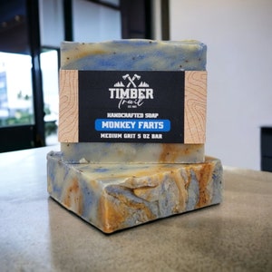 May include: Two bars of handmade soap with a blue and brown swirl pattern. The soap is labeled "Timber Trail" and "Monkey Farts" with a medium grit and 5 oz bar size.