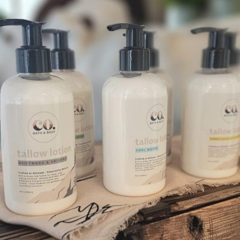 May include: Several white lotion bottles with black pumps are arranged on a wooden surface. The labels read "tallow lotion" with scent names like "Driftwood & Orchids" and "Cool Water". The bottles are from "Co. Bath & Body".
