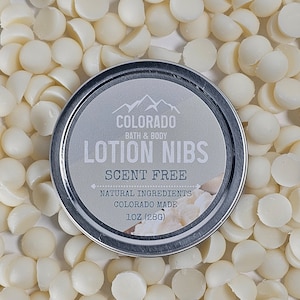 May include: A silver tin with a white label that reads "Colorado Bath & Body Lotion Nibs Scent Free Natural Ingredients Colorado Made 1 oz (28g)". The tin is surrounded by white lotion nibs.