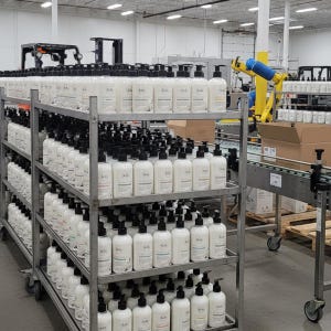 May include: Rows of white lotion bottles with black pump dispensers are stacked on metal shelves in a warehouse. The bottles have labels with minimalist designs. A forklift and robotic arm are visible in the background, suggesting a production or packaging facility.
