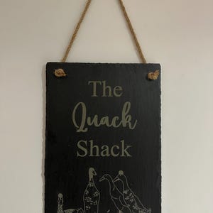 Op de afbeelding: Een zwart leisteen bord met een touw om op te hangen. Het bord heeft de tekst "The Quack Shack" in witte letters en toont een tekening van eenden.