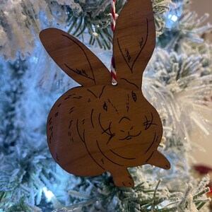 以下が含まれることがあります： 茶色の毛皮のウサギの形をした木製のクリスマスオーナメント。オーナメントは雪が積もったクリスマスツリーの赤と白の紐にぶら下がっています。