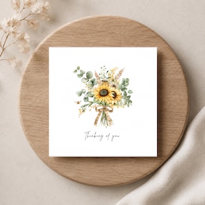 Può includere: Un biglietto d'auguri quadrato con un'illustrazione ad acquerello di un bouquet di girasoli gialli, foglie di eucalipto verdi ed erbe essiccate legate con un nastro a righe beige. Il testo Thinking of you è stampato in nero sotto il bouquet.