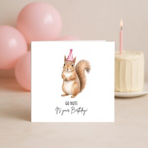 May include: A square white greeting card featuring a brown squirrel illustration wearing a pink polka-dot party hat. Below the squirrel, the text reads "GO NUTS It's your Birthday!" in black lettering. The card is centred against a neutral background.