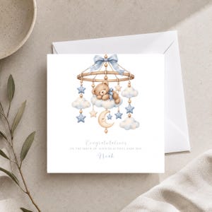 Peut inclure: Une carte de vœux carrée avec une illustration aquarelle d'un mobile pour bébé représentant un ours en peluche endormi, des nuages, des étoiles et un croissant de lune dans des tons bleu et beige. Le texte indique : Congratulations on the birth of your beautiful baby boy Noah.